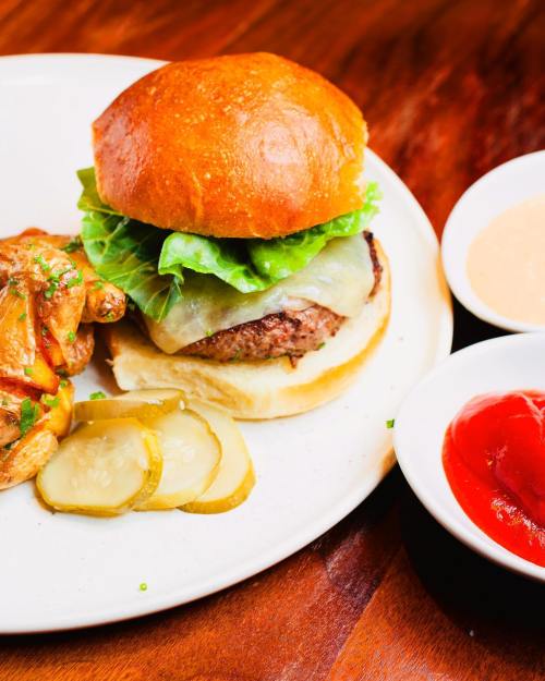 A juicy burger with lettuce and cheese on a bun, served with pickles, chicken wings, and dipping sauces.