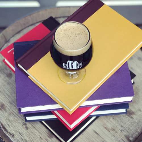 A glass of dark beer sits atop a stack of colorful books on a wooden surface.