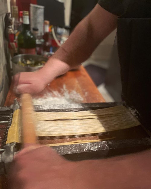 A person's hands rolling out pasta dough with a pasta machine on a wooden countertop, surrounded by flour and kitchen items.