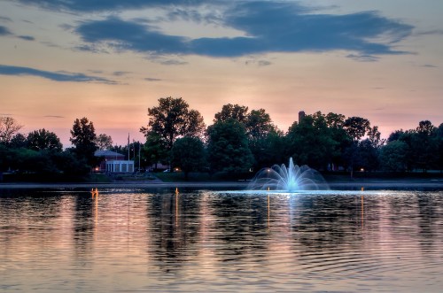 A serene lake at sunset, featuring a fountain and trees reflecting in the water under a colorful sky.