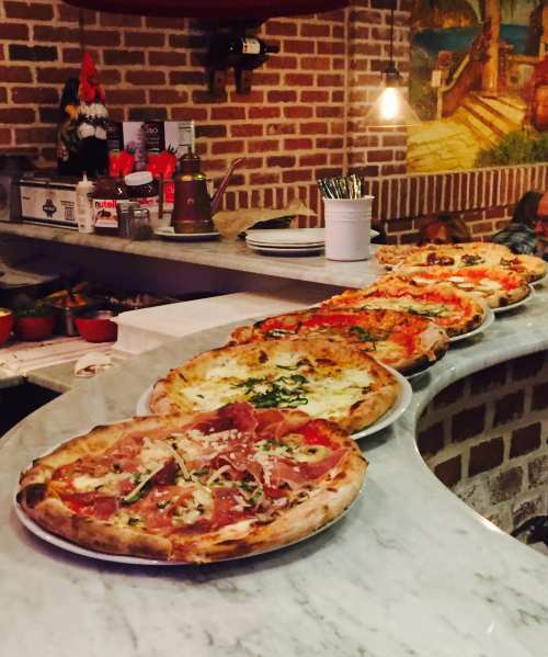 A variety of pizzas displayed on a marble counter in a cozy restaurant with a brick wall backdrop.