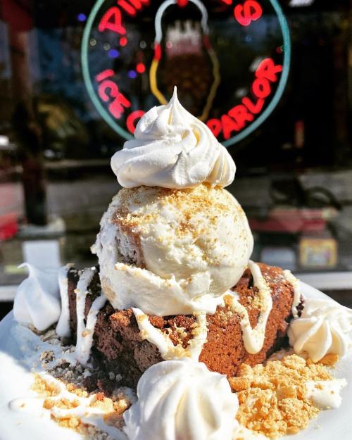 A decadent brownie topped with vanilla ice cream, whipped cream, and drizzled with sauce, set against an ice cream parlor backdrop.