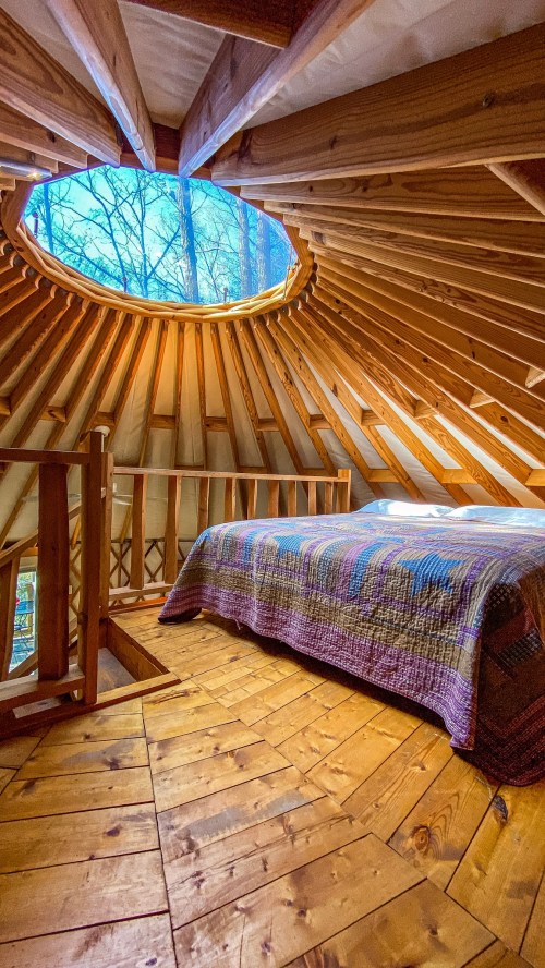 Cozy interior of a wooden yurt with a circular skylight and a bed, surrounded by natural wood beams and flooring.