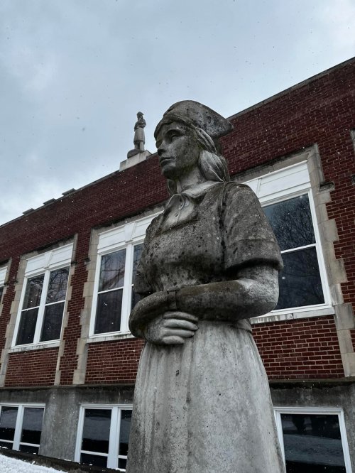 A weathered statue of a woman in a dress and cap stands in front of a brick building, with a cloudy sky above.