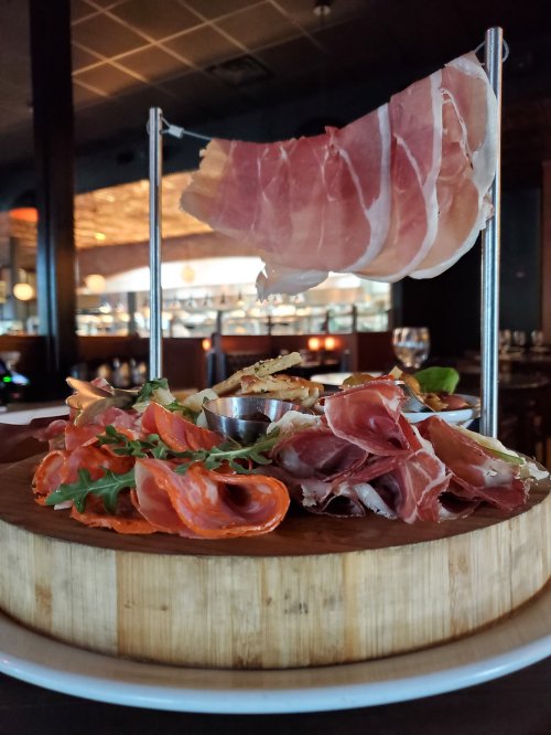 A wooden platter with assorted cured meats, garnished with greens and a small bowl of sauce, displayed elegantly.