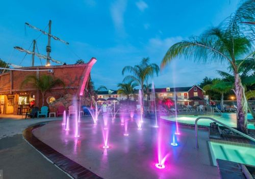 Colorful fountains illuminate a pool area with palm trees and a pirate ship structure under a twilight sky.
