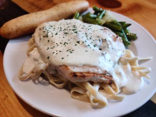 A plate of fettuccine topped with grilled chicken and creamy sauce, served with green beans and a breadstick.
