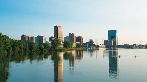 City skyline reflecting on calm water, surrounded by greenery under a clear blue sky.