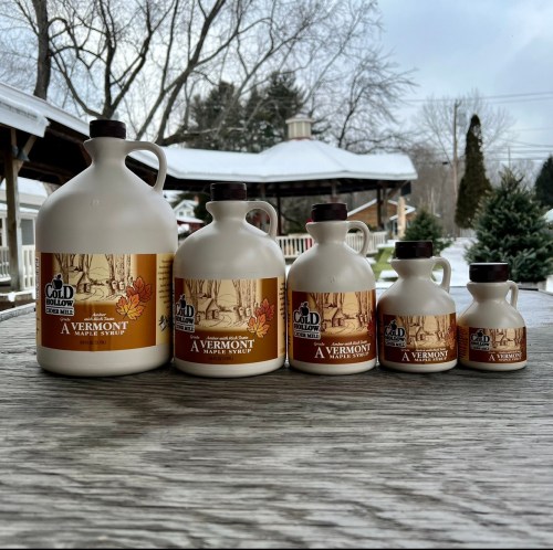 Five glass jugs of Vermont maple syrup in various sizes, set on a wooden table with a snowy background.