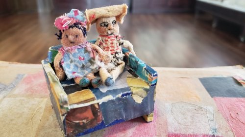 Two handmade dolls sit together on a small, colorful couch, surrounded by a textured background.
