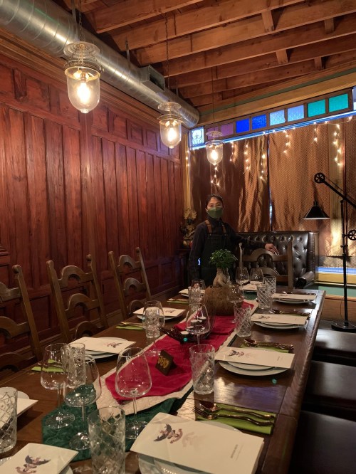 A cozy dining room with wooden walls, a long table set for a meal, and a person wearing a mask standing nearby.