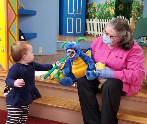 A toddler interacts with a woman in a pink shirt, holding a plush dragon toy in a colorful playroom.
