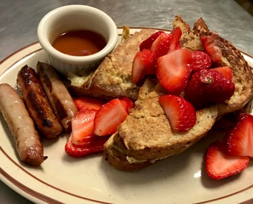 A plate of French toast topped with strawberries, served with sausage links and a small bowl of syrup.