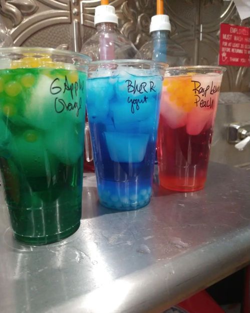Three colorful drinks in clear cups: green with orange, blue with yogurt, and red with peach, all with ice and toppings.