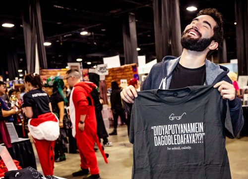 A smiling man holds up a humorous t-shirt at a convention, surrounded by colorful booths and attendees in costumes.