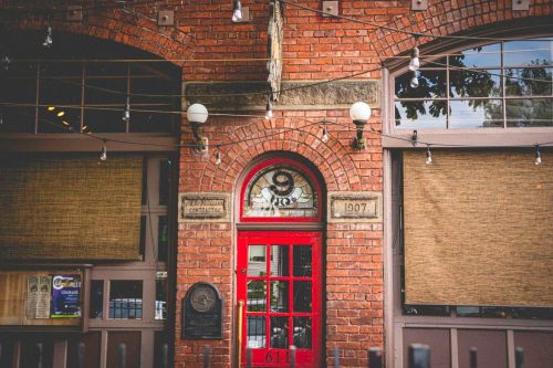 A red door with a decorative arch, surrounded by brick walls and large windows, featuring hanging lights and shades.