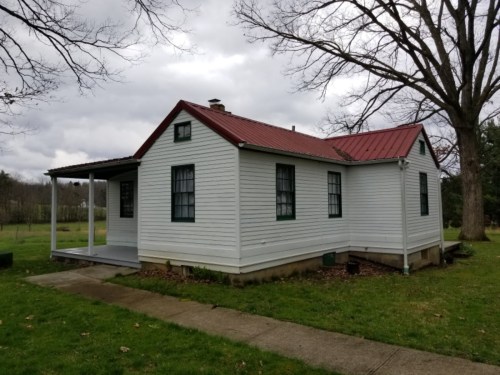 A small white house with a red metal roof, surrounded by grass and trees, under a cloudy sky.