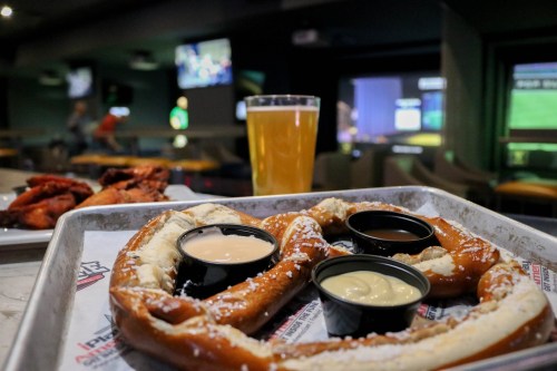 A large pretzel with dipping sauces, a pint of beer, and chicken wings on a tray in a sports bar setting.