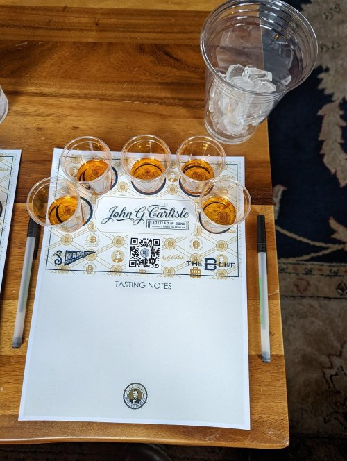 A wooden table with tasting notes, six whiskey samples in glasses, and a cup of ice.