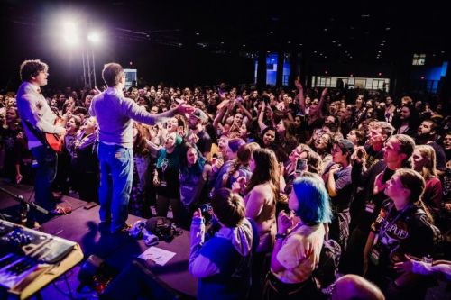 A lively concert scene with a crowd cheering for two performers on stage, surrounded by colorful lights and excitement.