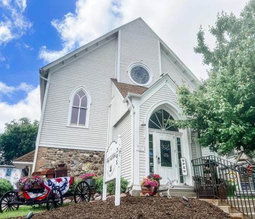 A charming white church with a stone base, featuring a round window and surrounded by colorful flowers and greenery.