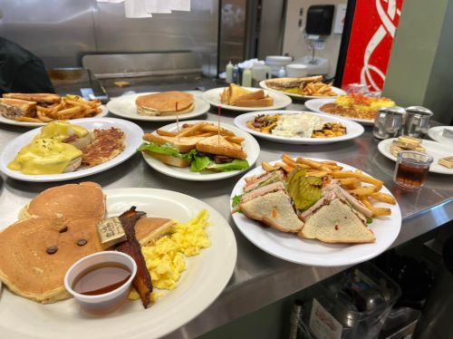 A variety of breakfast and lunch dishes on plates, including pancakes, sandwiches, fries, and eggs, on a diner table.