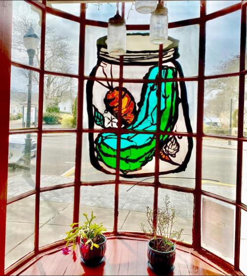 Stained glass window featuring a pickle jar with a pickle and a flower, framed by a cozy café setting.