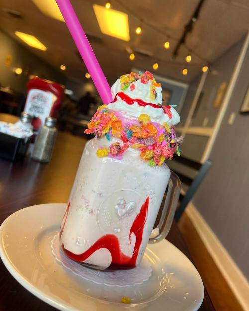 A colorful milkshake topped with whipped cream, sprinkles, and a cherry, served in a mason jar with a pink straw.