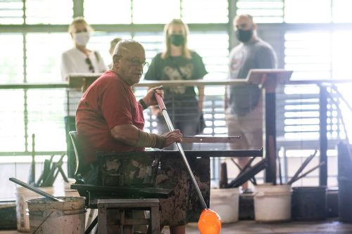 A glassblower shapes molten glass while onlookers watch from a distance, all wearing masks in a workshop setting.