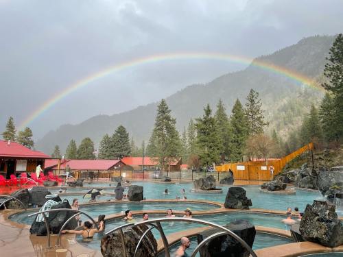 A scenic hot spring with people relaxing, surrounded by mountains and a vibrant rainbow in the sky.