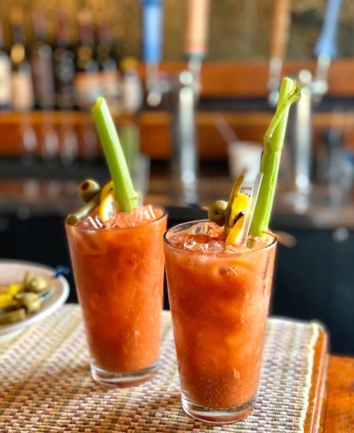 Two tall glasses of Bloody Mary cocktails garnished with celery, olives, and lemon slices, sitting on a bar counter.