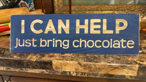 A wooden sign with blue background reads, "I CAN HELP just bring chocolate" in bold, light-colored letters.