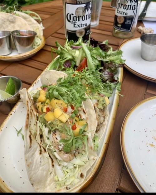 A plate of tacos topped with mango salsa, served with a side salad and drinks on a wooden table.