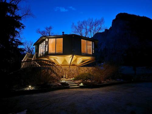 A modern, elevated house with large windows, surrounded by trees, illuminated at dusk against a mountain backdrop.