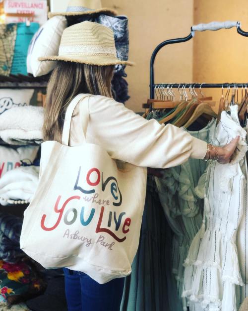 A person wearing a straw hat browses clothing on a rack, holding a tote bag that says "Love where You Live Asbury Park."