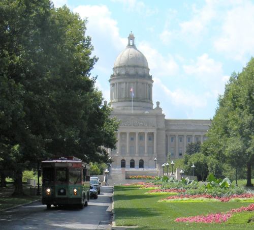 A historic building with a dome, surrounded by greenery and colorful flowers, with a trolley and cars in the foreground.
