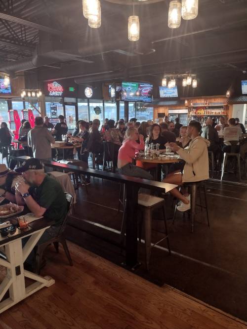 A lively restaurant scene with patrons dining and socializing, featuring bar seating and multiple TVs showing sports.
