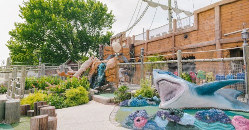 A colorful outdoor play area featuring a pirate ship, waterfall, and a large shark sculpture among lush greenery.