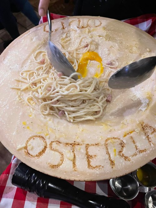 A large cheese wheel with spaghetti and an egg in the center, surrounded by flour, with "OSTERIA" written on it.