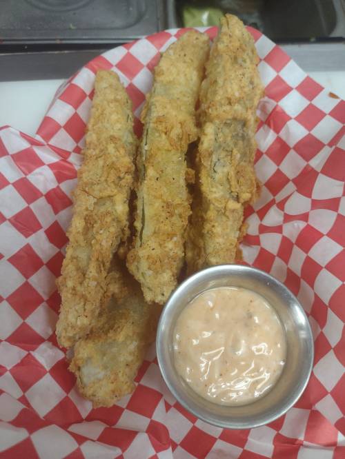 Three crispy fried pickles served with a small cup of dipping sauce on a red and white checkered paper.