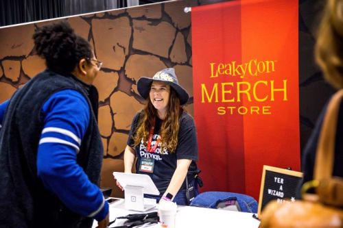 A smiling staff member at the LeakyCon merch store interacts with a visitor at a convention booth.