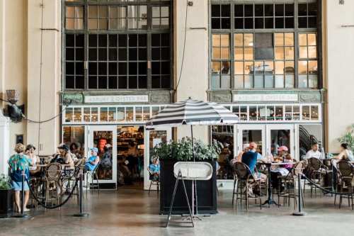 A bustling café with large windows, featuring patrons seated at tables and a striped umbrella in the center.