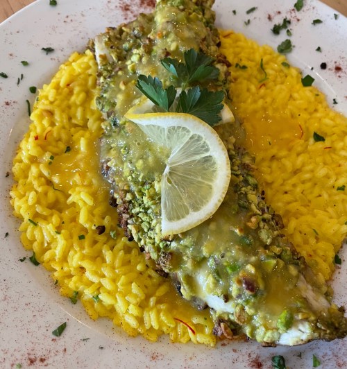 A plate of yellow saffron rice topped with a baked fish fillet, garnished with lemon slices and parsley.