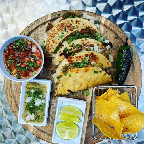 A wooden platter with tacos, salsa, guacamole, lime wedges, and tortilla chips, garnished with cilantro.