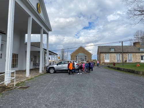 A group of people walks towards a parked car near a white building and a stone structure under a cloudy sky.