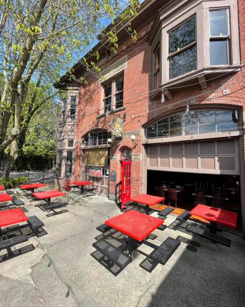 A brick building with large windows and a red door, featuring outdoor seating with red tables and chairs.
