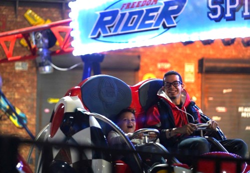 Two people, a man and a child, smile excitedly on a carnival ride at night, illuminated by colorful lights.
