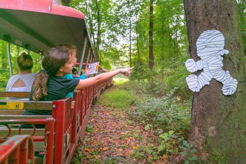 A child points at a paper figure on a tree while riding a small train through a wooded area.