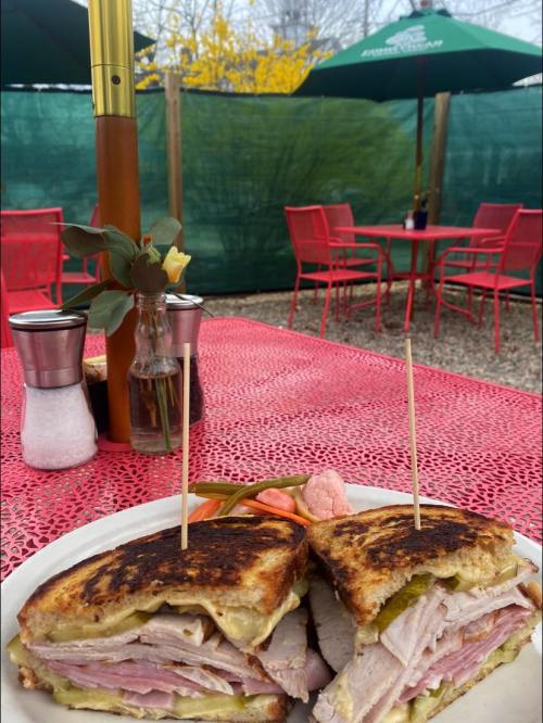 A close-up of a sandwich with layers of meat, served on a plate, with a colorful outdoor dining area in the background.