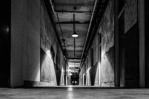 A dimly lit, long corridor with peeling walls and overhead lights, creating a moody, abandoned atmosphere.
